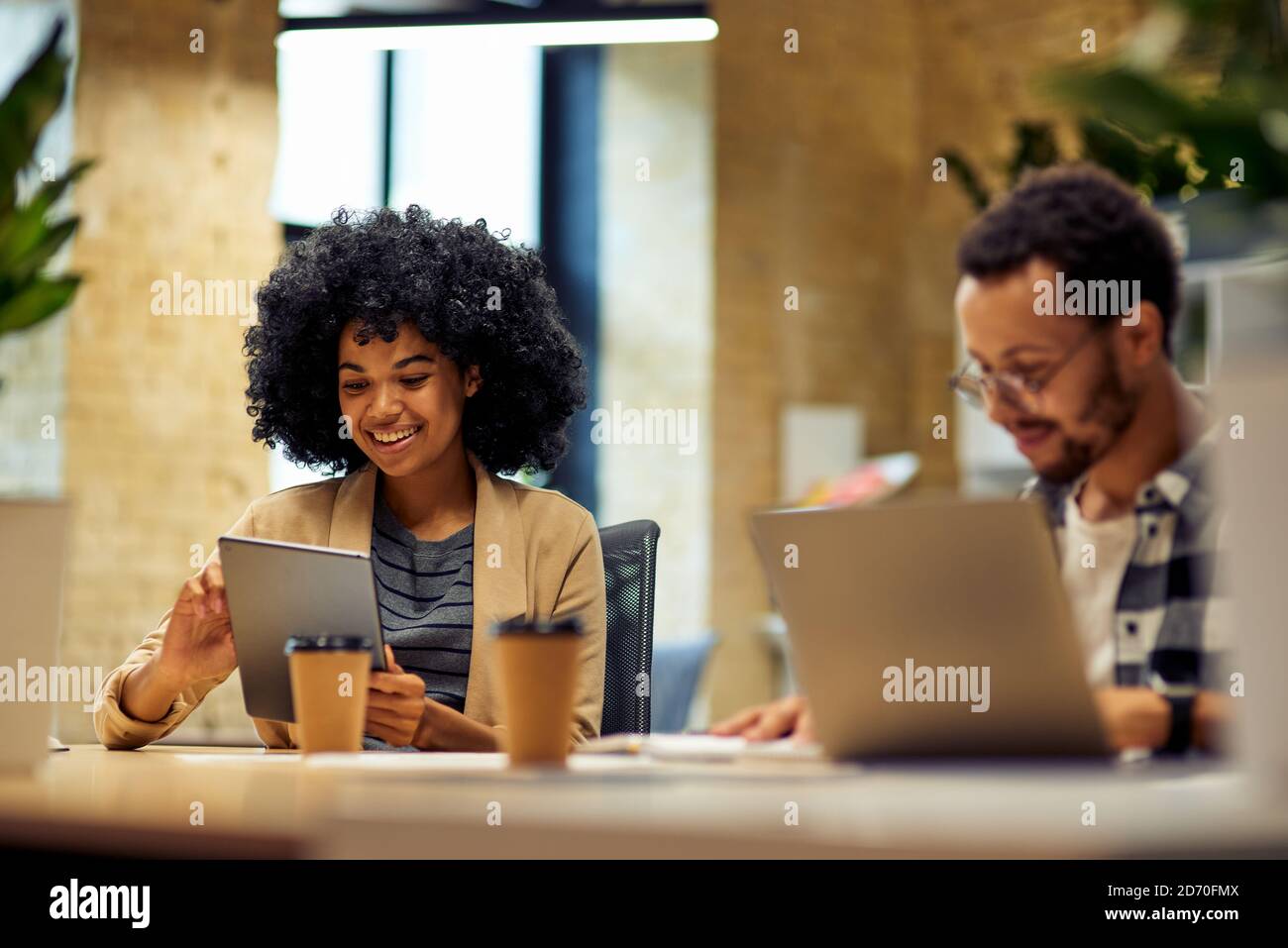 Two young happy multiracial business people sitting at the desk, using modern technologies while working together in coworking space. Office life, teamwork and business Stock Photo