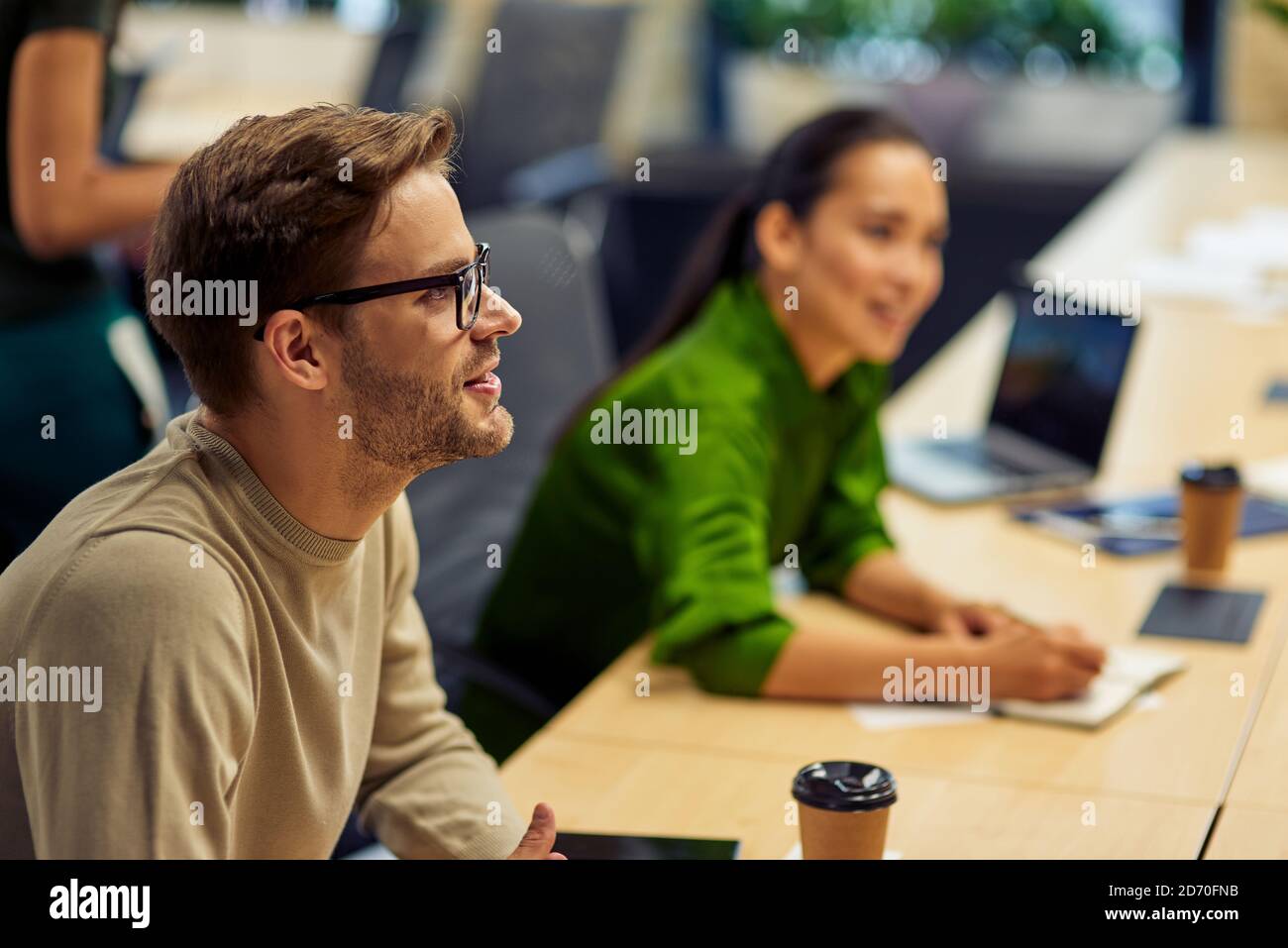 Side view of a young handsome man, male office worker sitting at desk in the modern office and looking aside while working with colleagues. Business people and office life, teamwork Stock Photo