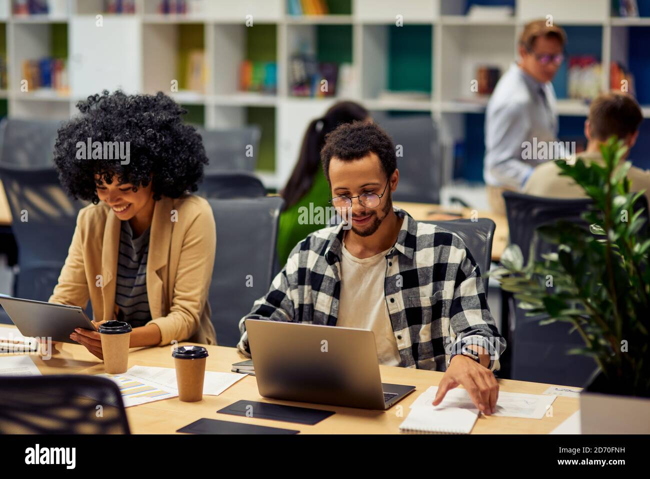 Two young diverse business colleagues using modern technologies while sitting at the desk and working together in coworking space. Office life, teamwork and business Stock Photo