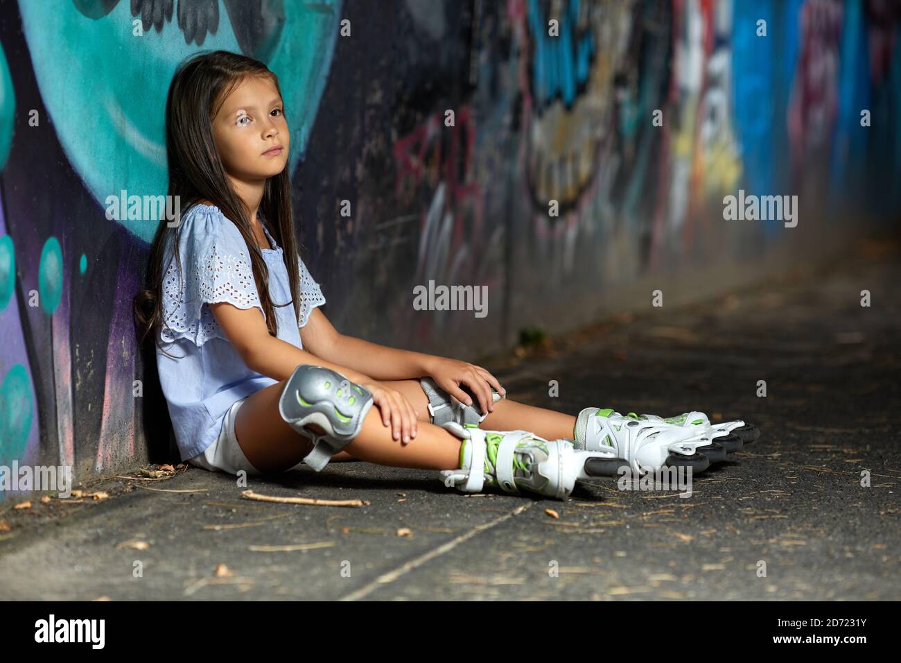 tired happy little child girl in roller skates is sitting after rollerskating in park in the