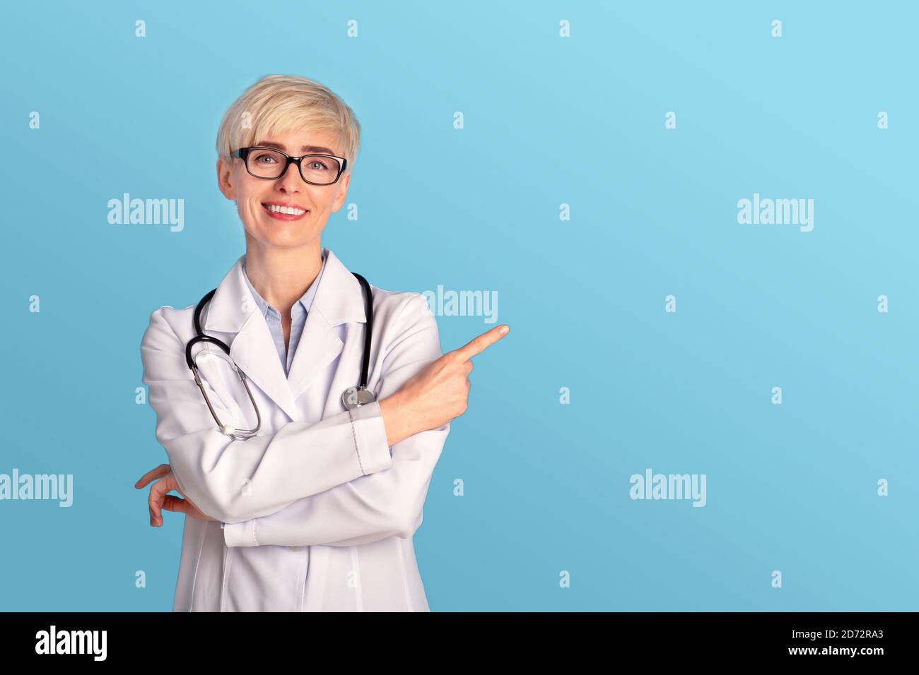 Smiling lady doctor in glasses, white coat with stethoscope points finger to free space for good treatment Stock Photo