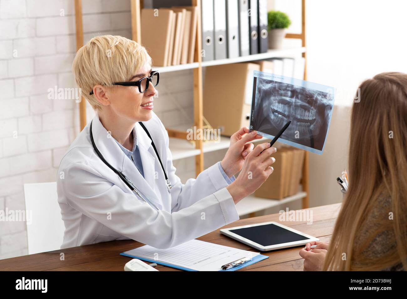 Adult lady doctor in white coat and glasses shows x-ray of patient jaw and teeth and diagnoses, sitting at table Stock Photo