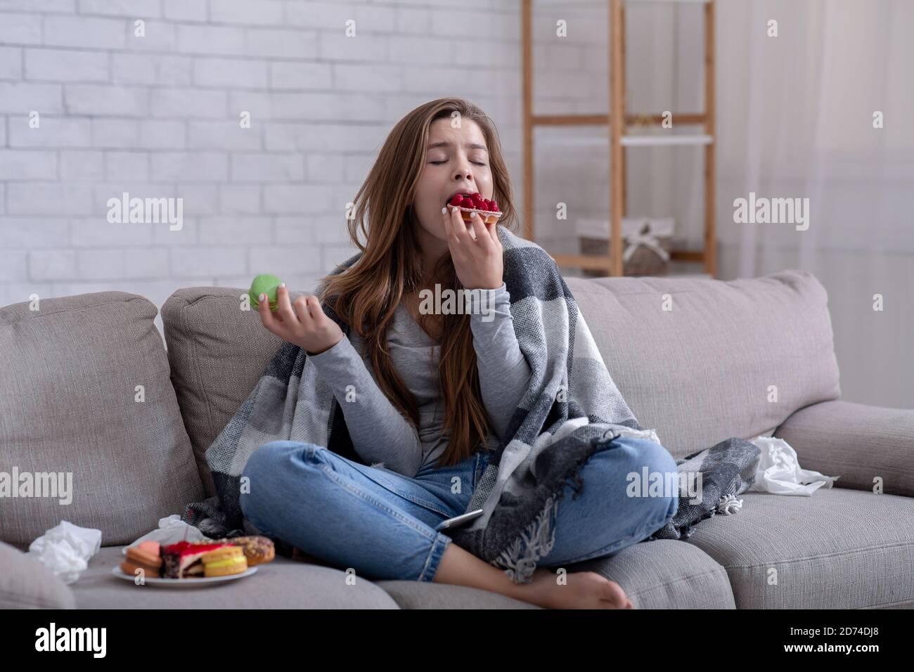 Sad young lady coping with depression or stress by eating sweets on sofa at home Stock Photo