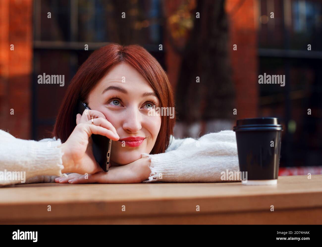 Woman leans on bar table at outdoor cafe terrace and speacks on a phone with funny emotional expression on her face. Stock Photo