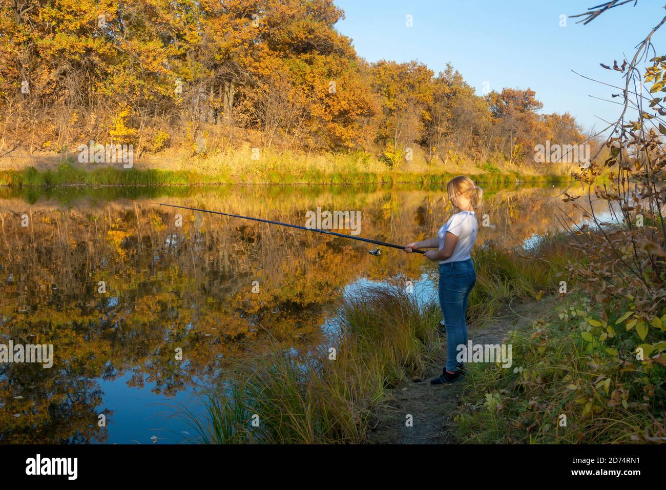 Young blonde woman is fishing with fishing rod in autumn on river. Fishing for carp, perch on lake or pond. Stock Photo