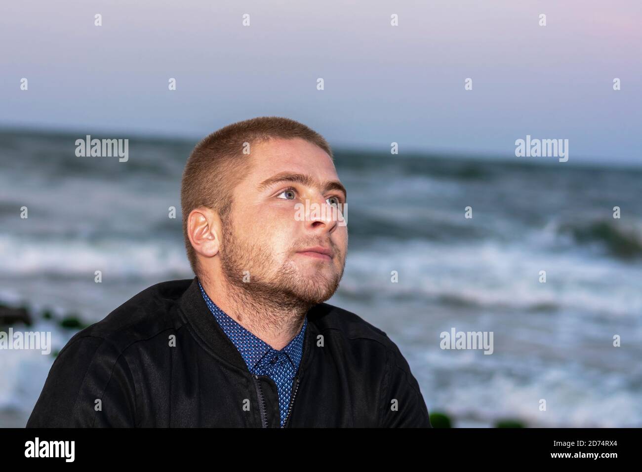 Portrait of young handsome man against background of sea in the evening. Look up, romantic mood. Stock Photo
