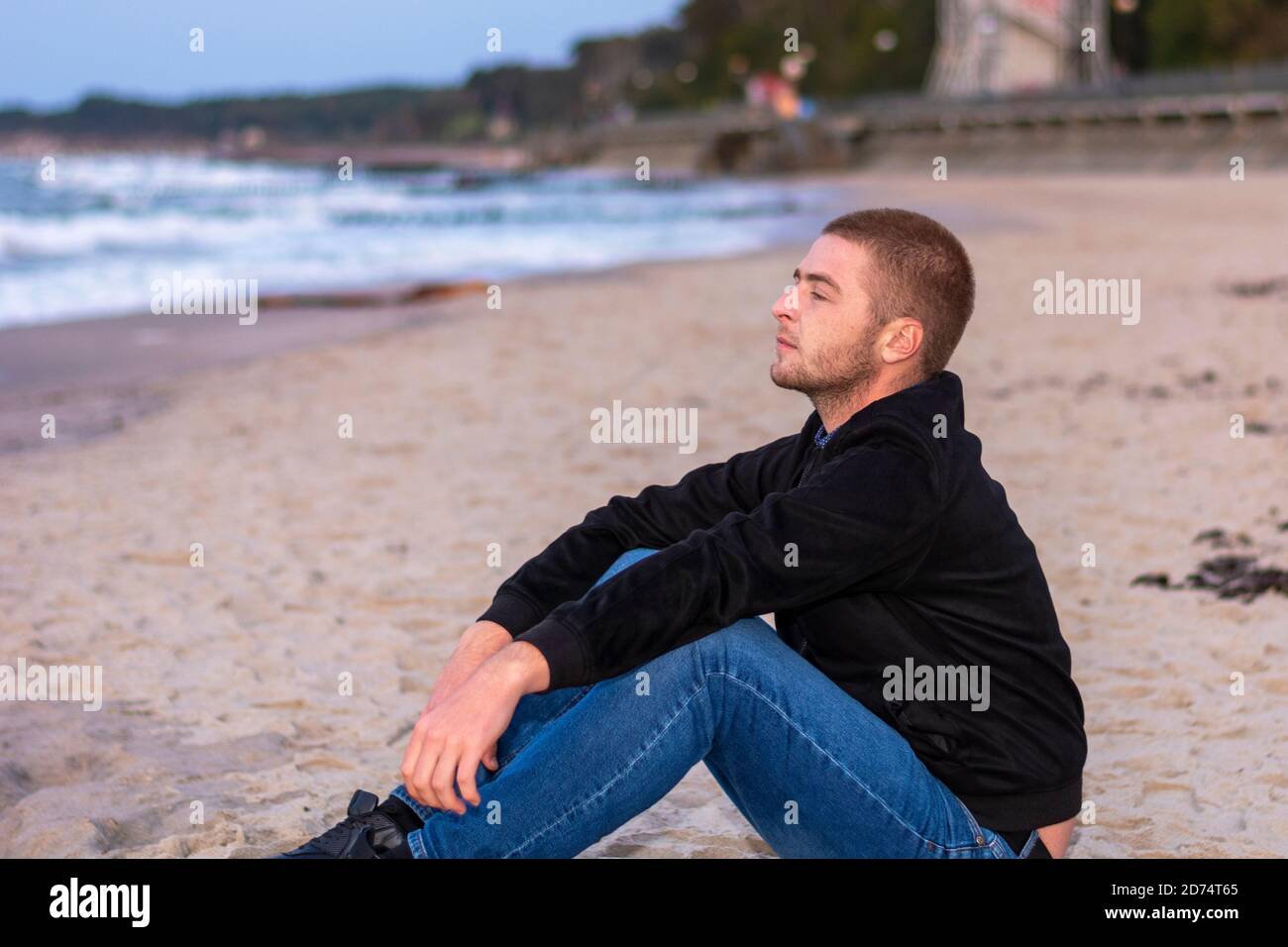 Young man sits in profile on seashore in evening and looks at water. Romantic mood, selective focus. Stock Photo