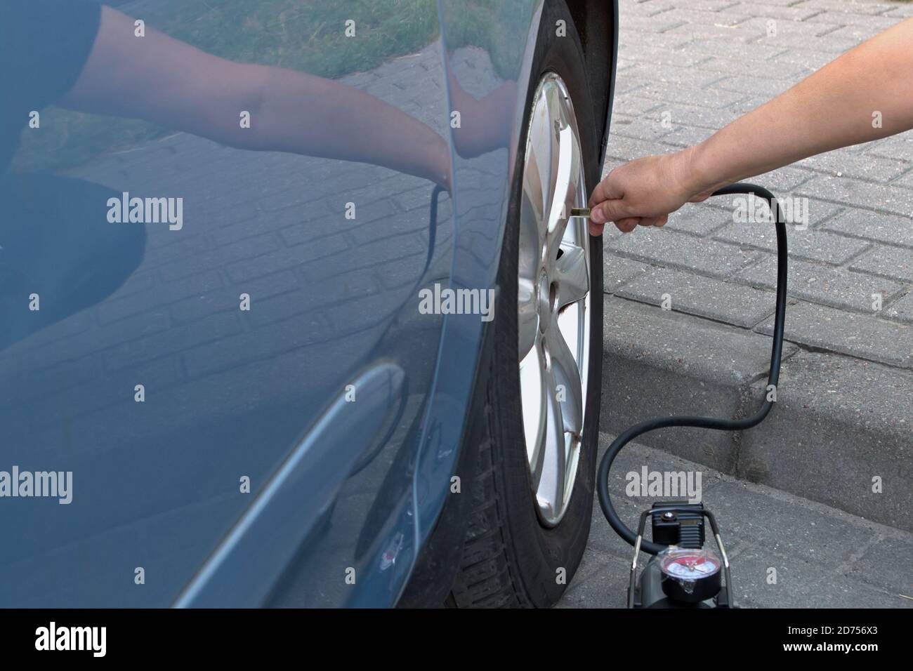 Woman squatting on sidewalk of gray paved street parking lot pumps  front wheel of blue car with black compressor. Concept: woman doing man's job Stock Photo