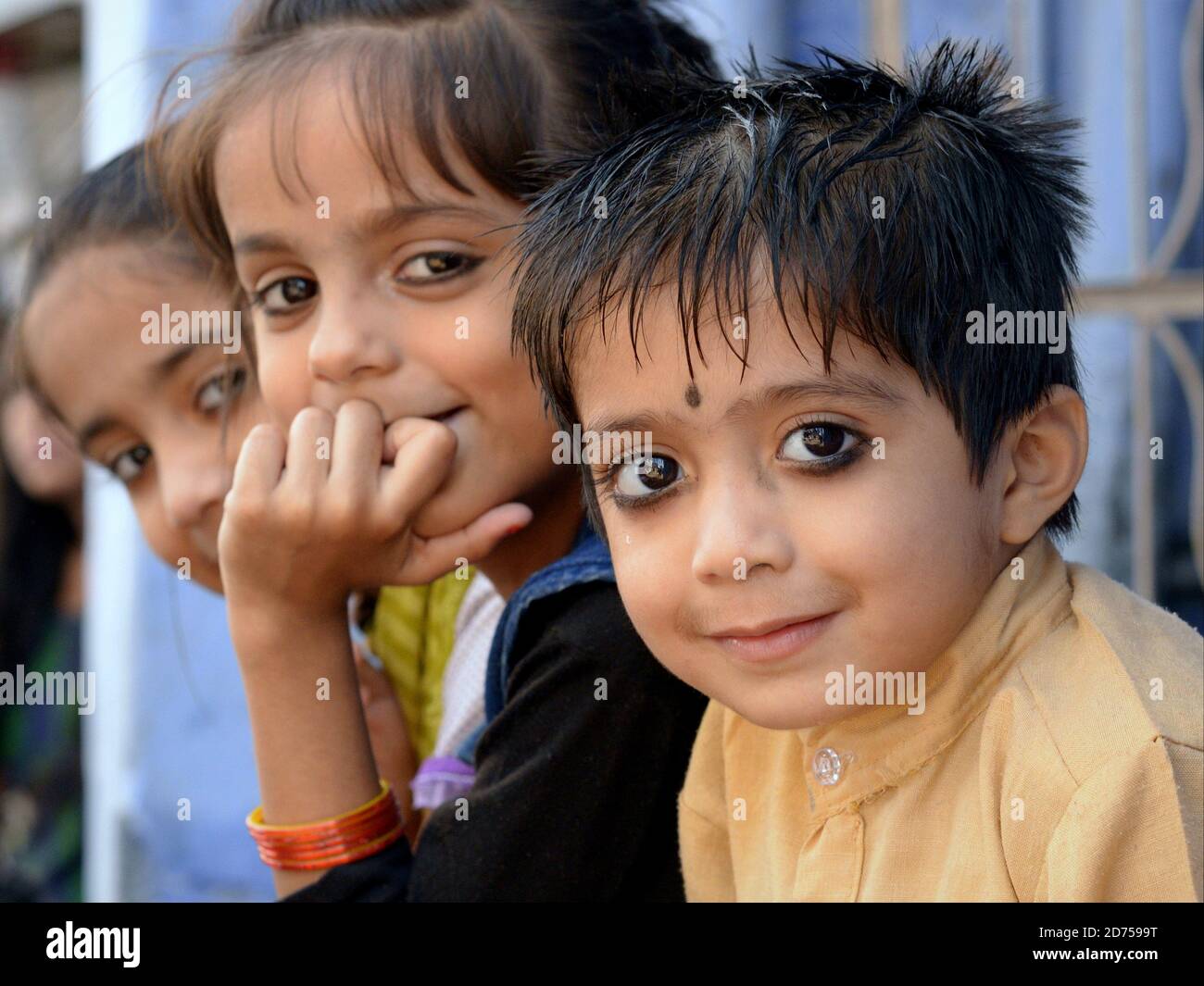 Three little Indian Rajasthani children with traditional kohl eyeliner sit in a row and pose for the camera. Stock Photo