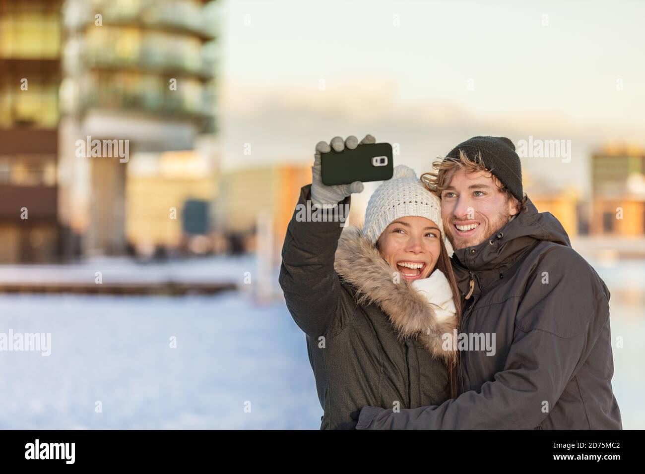 Selfie couple happy winter travel walk tourists taking photo with phone on city street panorama lifestyle. Asian woman, Caucasian man wearing knit Stock Photo