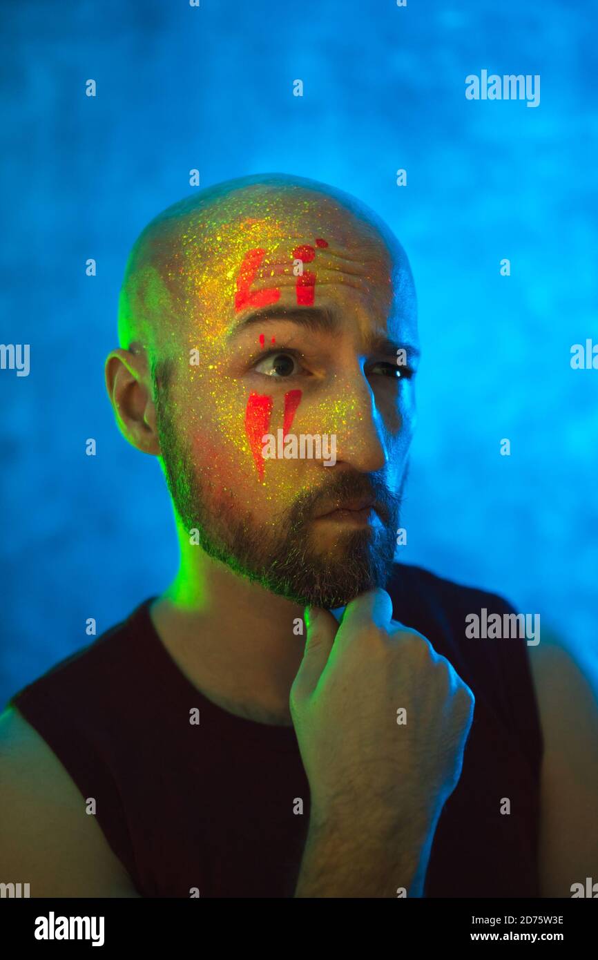 Pensive bald man with glowing fluorescent makeup on face in neon lighting. Ethnic, esoteric and art concept. Stock Photo