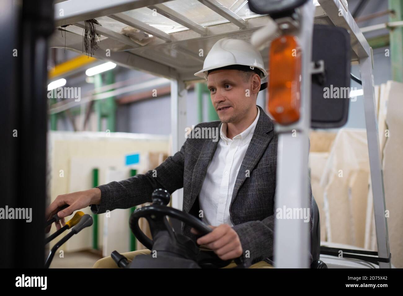 Elegant male employee in jacket and hardhat operating forklift while working in modern warehouse Stock Photo