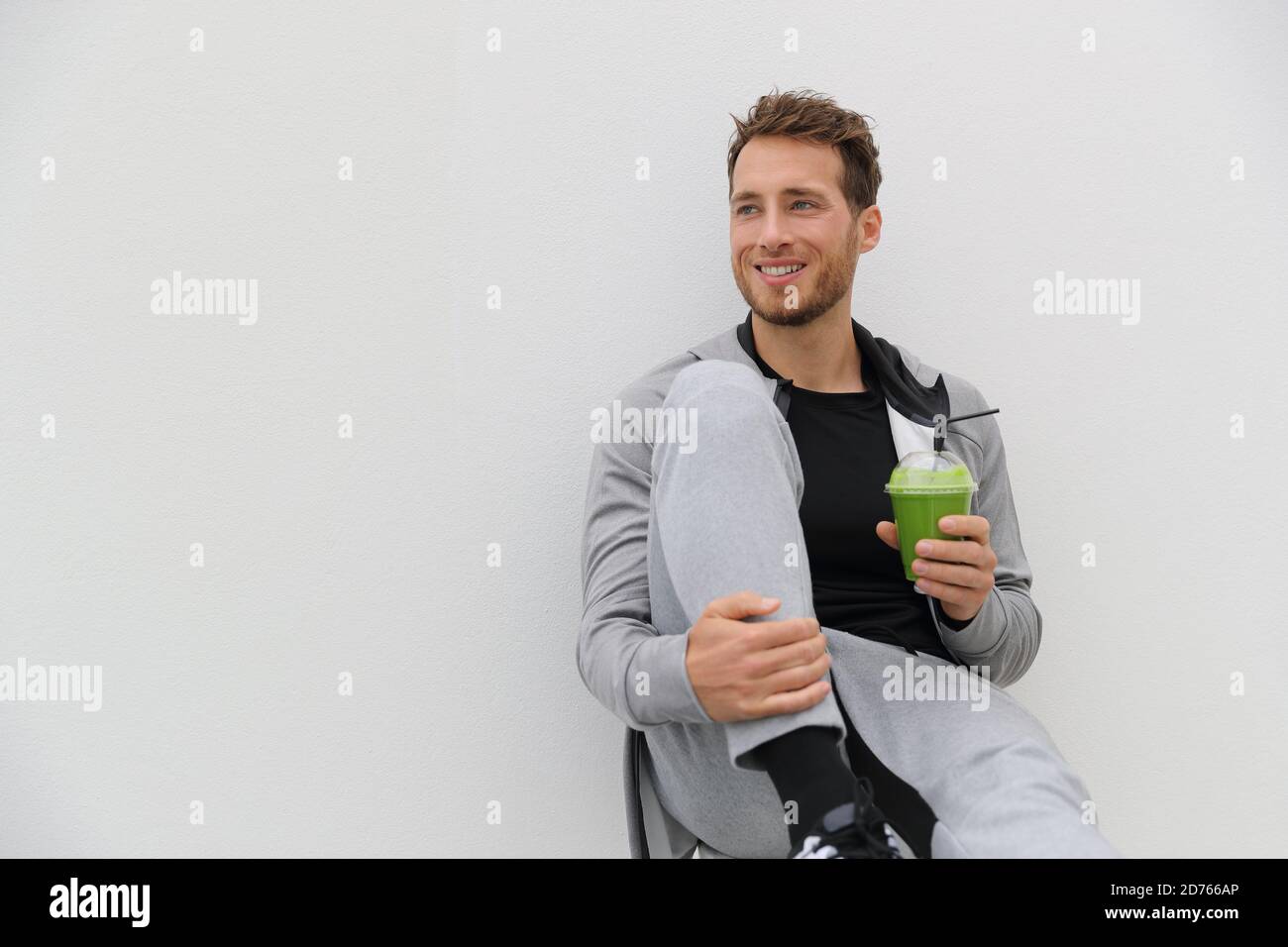 Healthy man drinking green smoothie post workout at gym. Sport athlete with spinach cold pressed juice plastic cup drink relaxing on white background Stock Photo