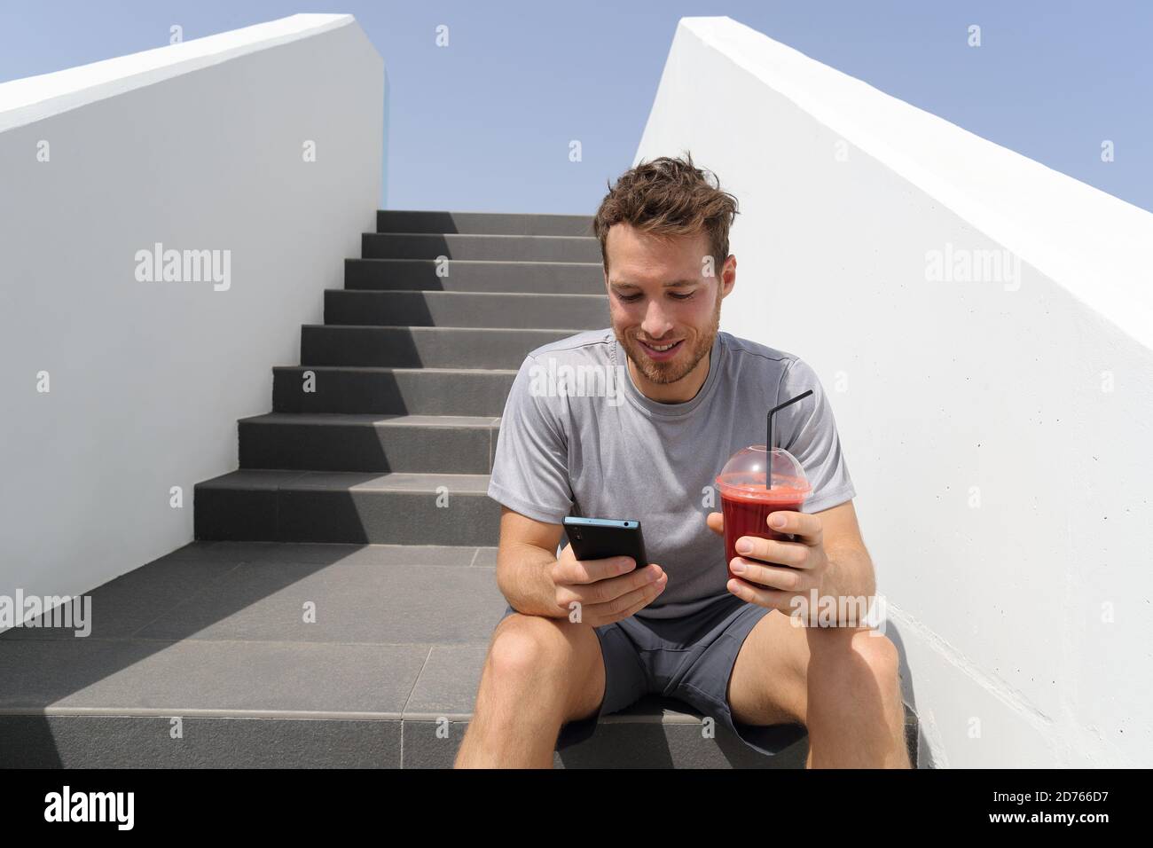 Healthy smoothie drinking man using phone app on jogging break with morning red beet juice sitting on stairs outdoor Stock Photo