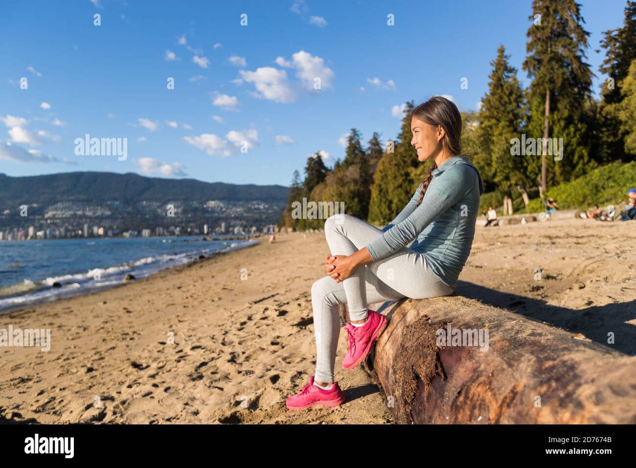 Vancouver urban lifestyle woman relaxing on Third Beach in Stanley Park, Vancouver, BC, Canada. Canadian Asian girl sitting on tree trunk at popular Stock Photo