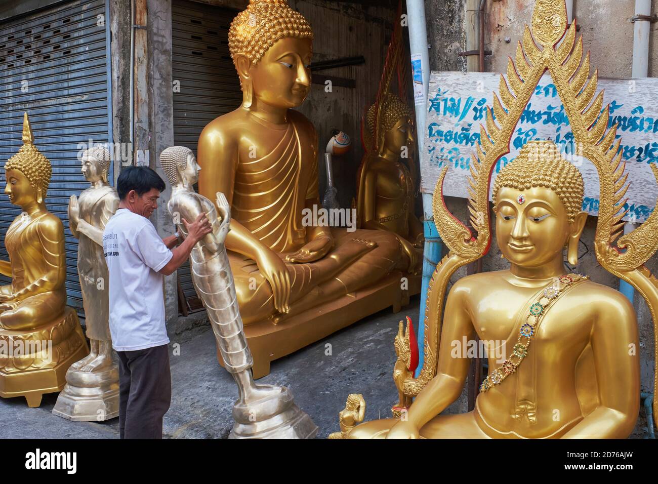Next to several finished Buddha statues, an employee of a Buddha factory moves one statue for some final work; Bamrung Muang Rd., Bangkok, Thailand Stock Photo
