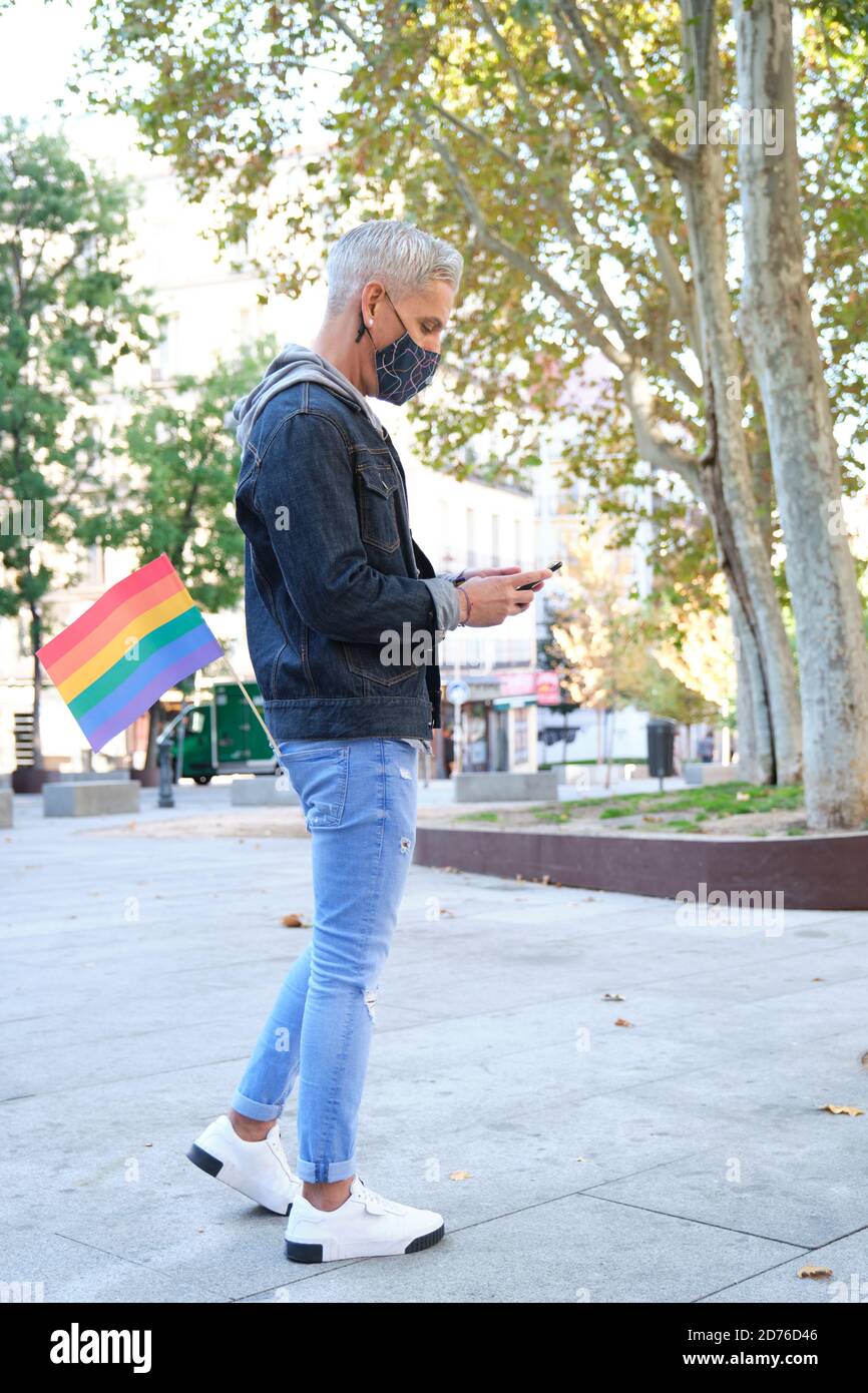 Young caucasian homosexual man wearing face mask with an LGBT flag in his pocket chatting on his smartphone. LGBT pride celebration in pandemic times. Stock Photo