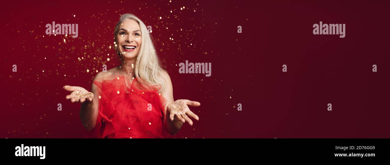 Happy holidays with excited senior woman throwing glitters in studio. Caucasian senior woman playing with golden star glitters over red background. Stock Photo