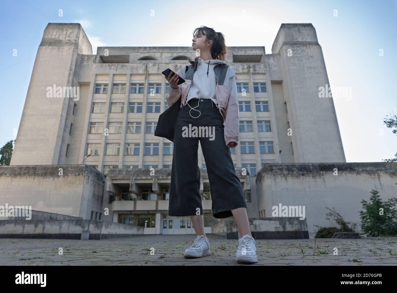 Young trendy teenage student standing in a street waiting in a low angle view against a high-rise office block or college listening to music on her mo Stock Photo