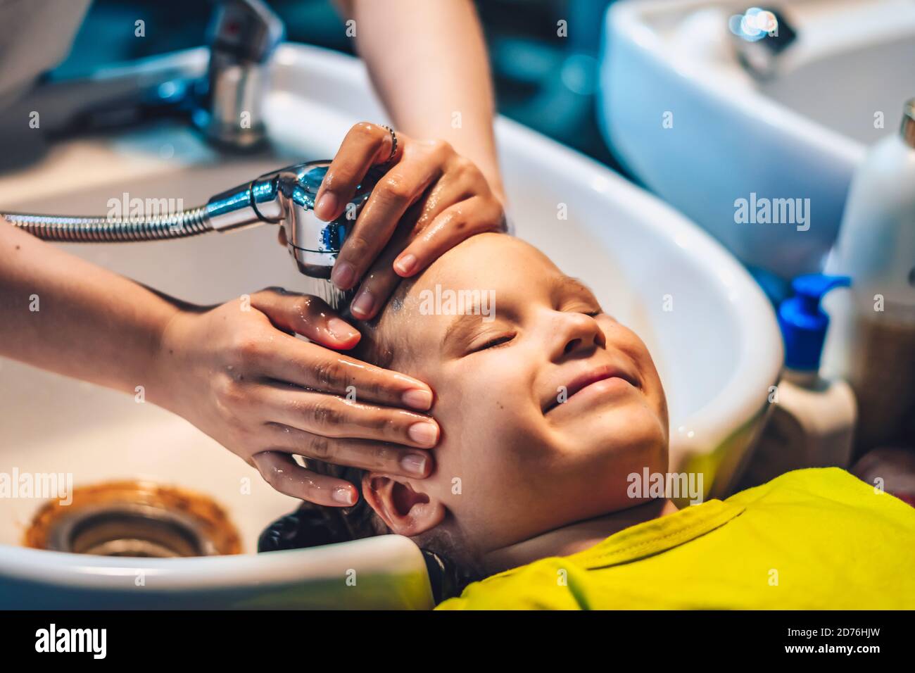 Wet Head Blond little Boy lie leaning on over sink. Woman female hands. Hair wash shower water stream, beauty hairdressing salon Work. Child enjoy Stock Photo