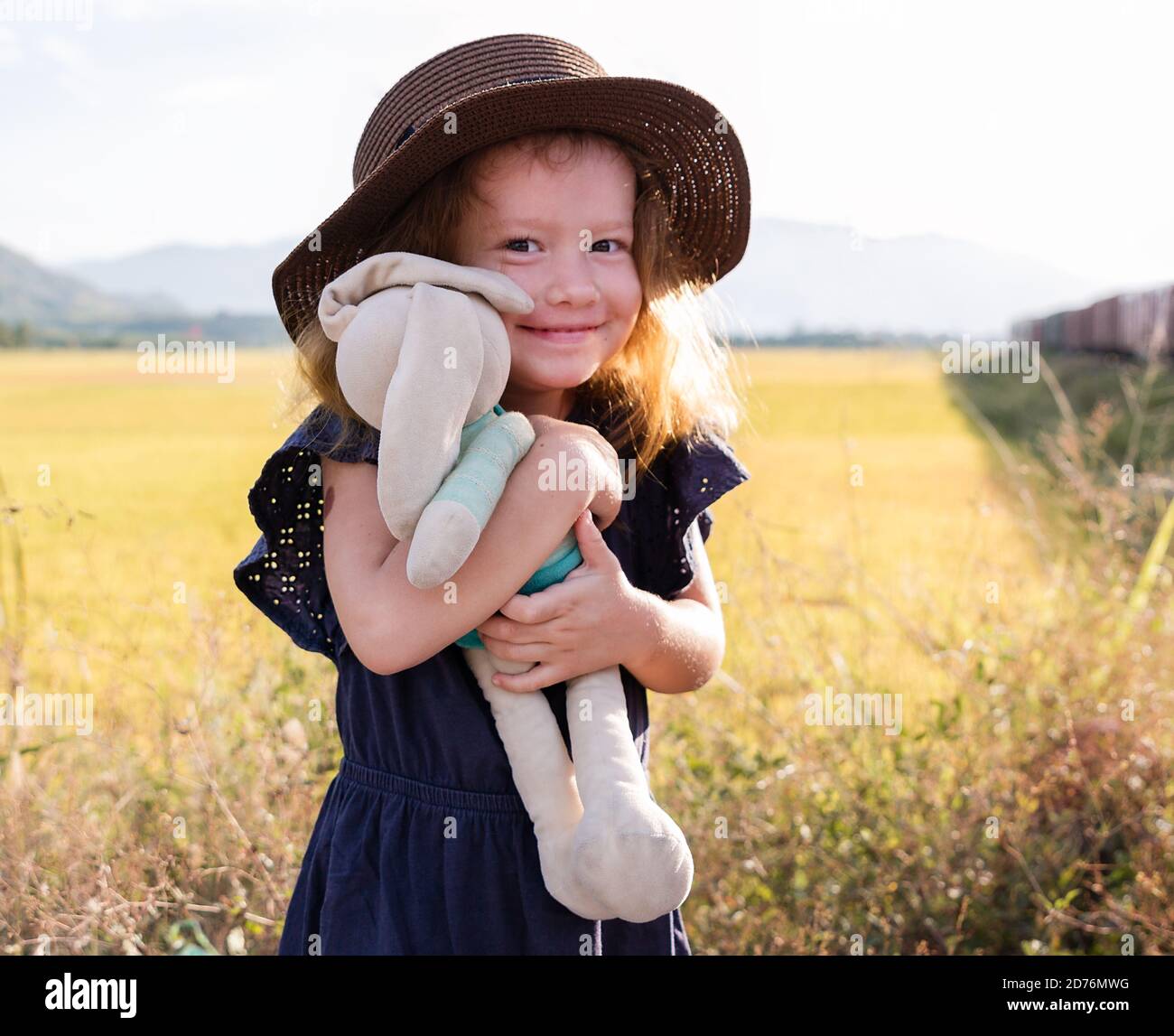 Child girl in straw hat. Cute kid with soft rabbit toy looking at nature lanscape background. Adventure travel concept in retro style Stock Photo