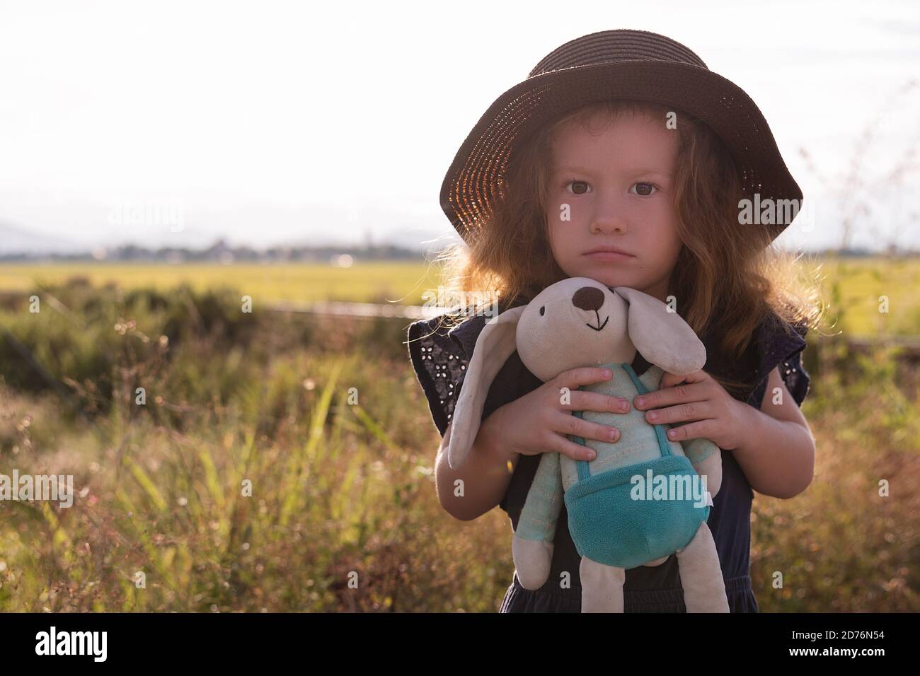 Child girl in straw hat. Cute kid with soft rabbit toy looking at nature lanscape background. Adventure travel concept in retro style Stock Photo