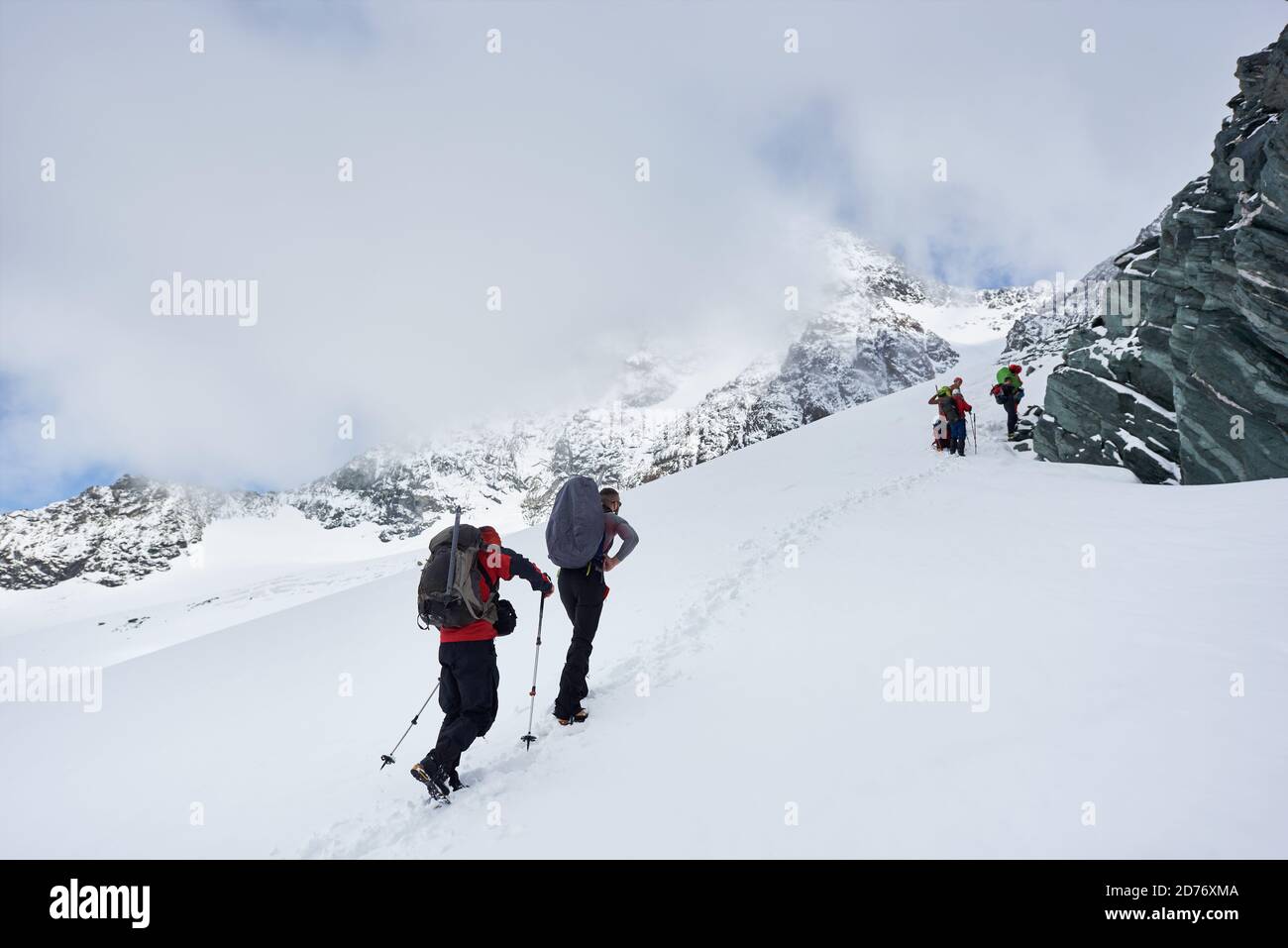 Male travelers with backpacks walking uphill in winter mountains. Group of alpinists with trekking sticks walking through snow and heading to mountain Stock Photo