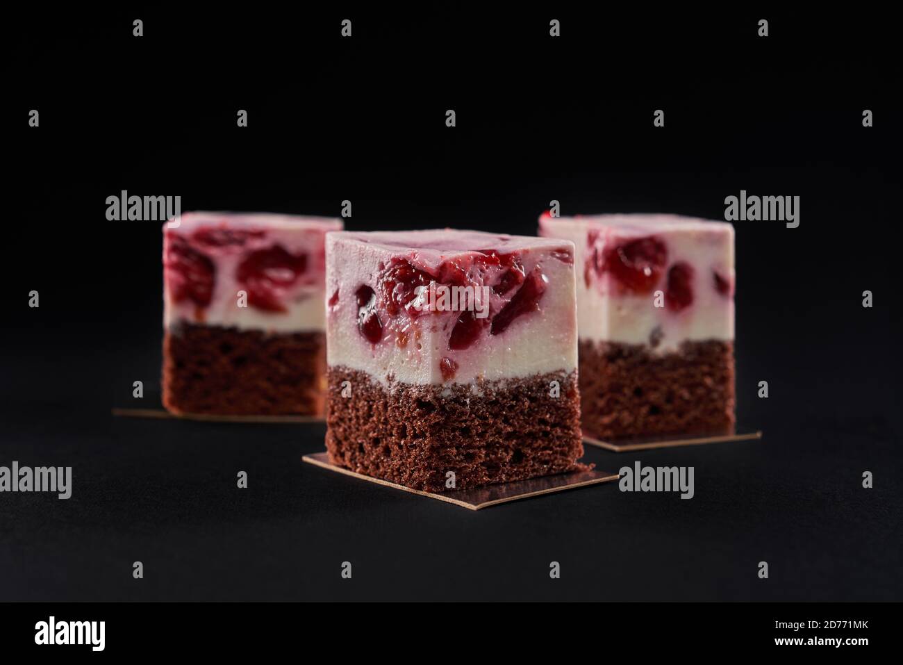 Front view of three delicious fresh pieces of cake made with brown biscuit and mousse pink layer with red jelly cherries. Tasty sweet dessert isolated on black background at restaurant. Stock Photo