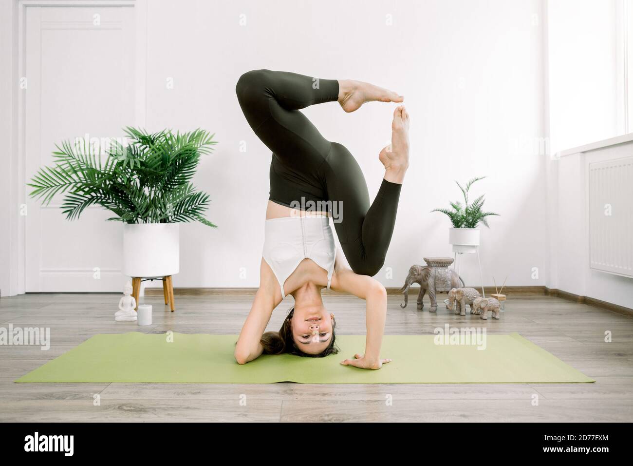 Full length indoor shot of young Caucasian woman practicing yoga while standing on head in sirsasana yoga pose with legs apart. Yoga, harmony and Stock Photo