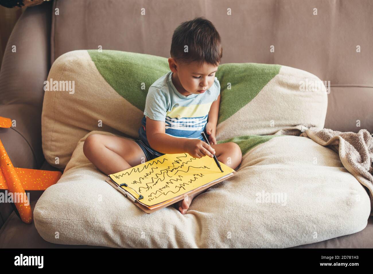 Caucasian boy drawing something on a yellow paper with a marker while sitting in bed on pillows Stock Photo