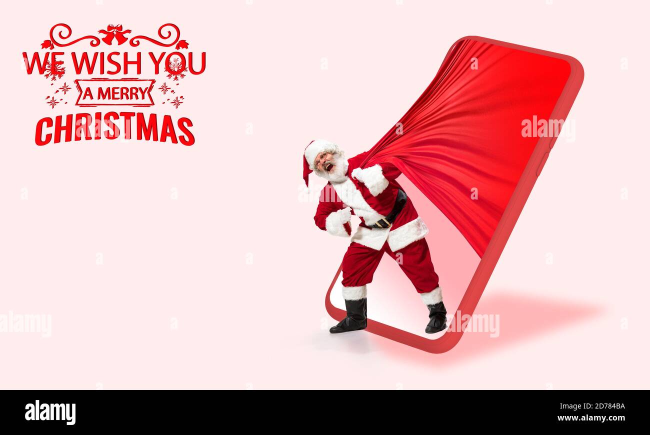 Emotional Santa Claus greeting with New Year 2021 and Christmas. Caucasian man in traditional costume. Concept of holidays and winter mood. Flyer with cpyspace. Walking throught the phone screen. Stock Photo