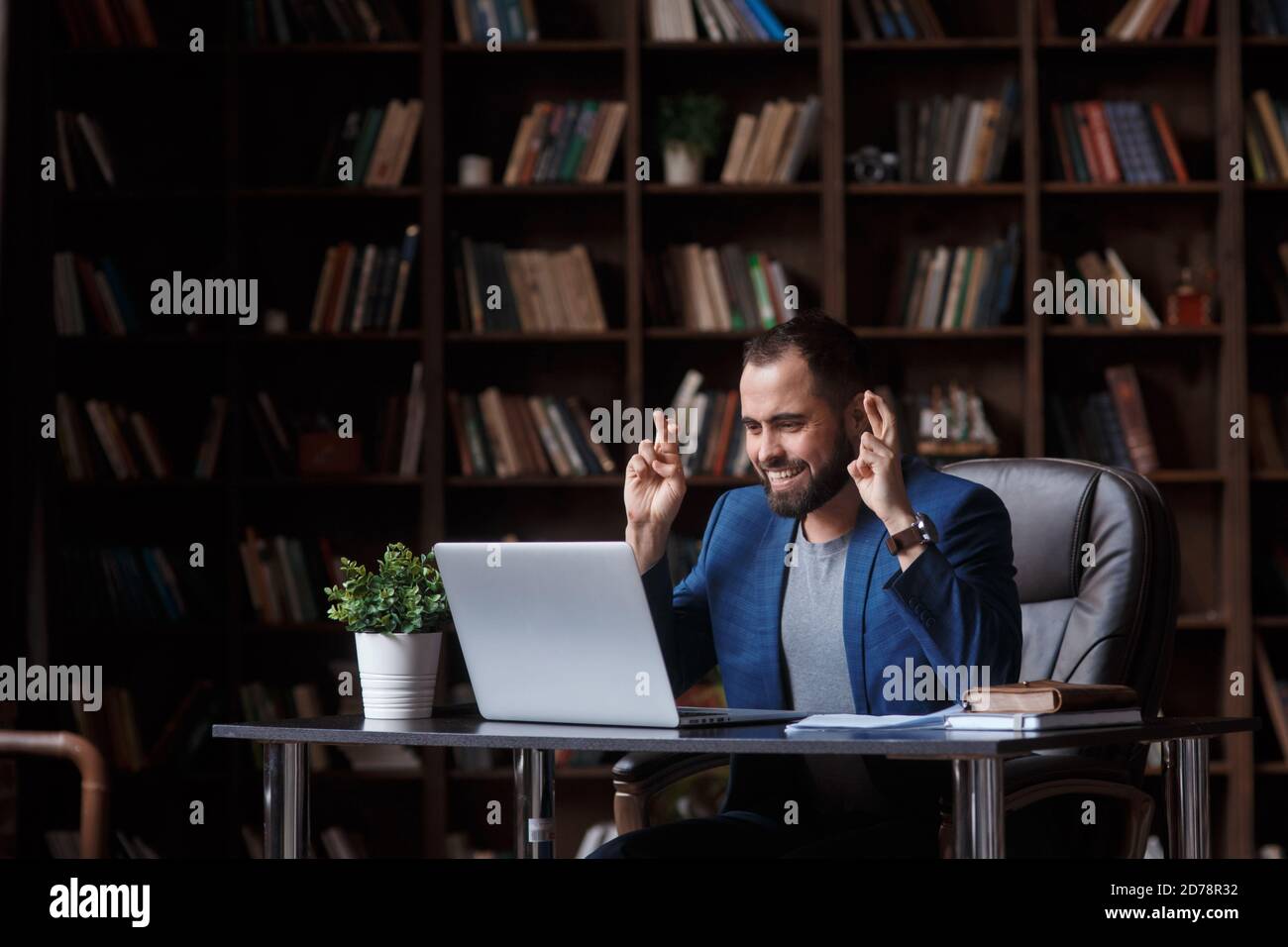A young bearded businessman in the library office in a blue suit looks at the laptop screen and crosses his fingers. concept of waiting for the Stock Photo