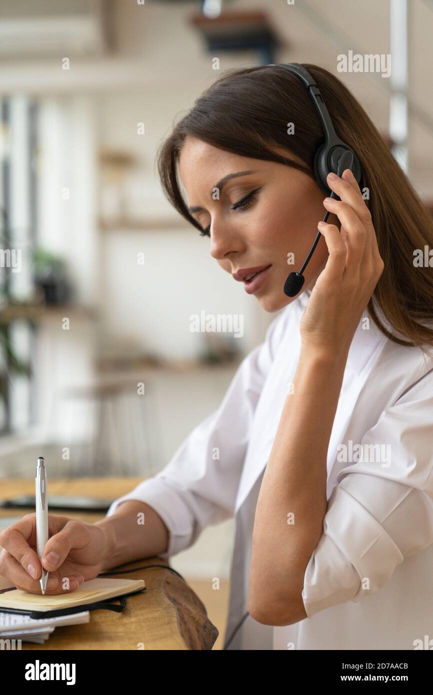Focused woman consultant in shirt wear headphones communicating with clients, talking on video conference, makes a note in notebook, remote work from Stock Photo