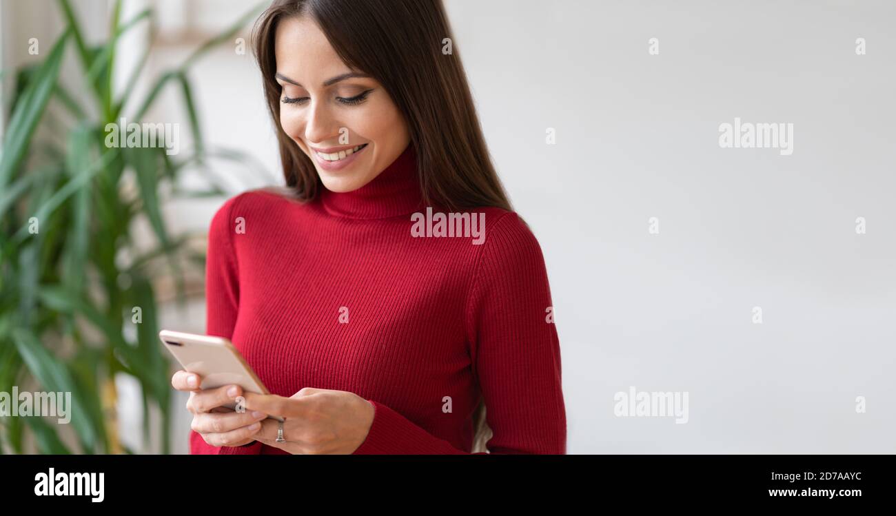 Satisfied cheerful woman wear red turtleneck holding mobile phone, makes shopping online, surfing internet received message from friend chatting about Stock Photo