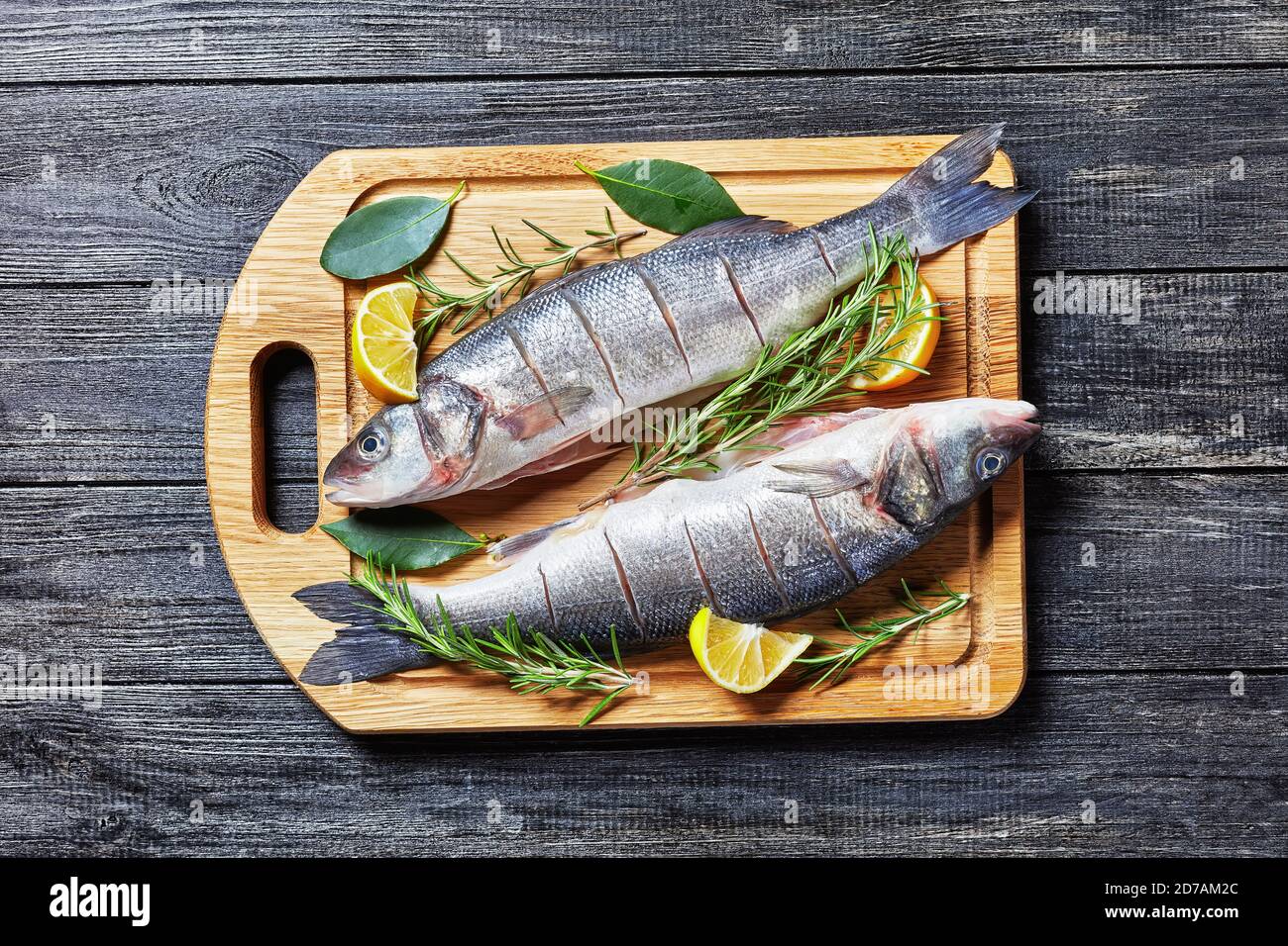 Sea Bass fresh raw seafood gutted and descaled on a cutting board with fresh rosemary, lemon wedges and bay leaf on a cutting board on a dark wooden b Stock Photo