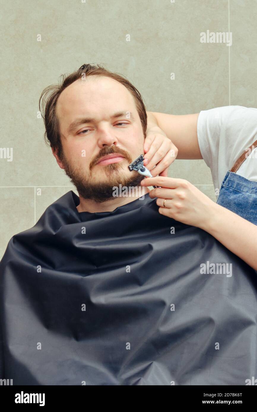 A woman shaves a man's beard with a safety razor. Concept of closed hairdressers and barbershops in isolation from the epidemic of the coronavirus Stock Photo