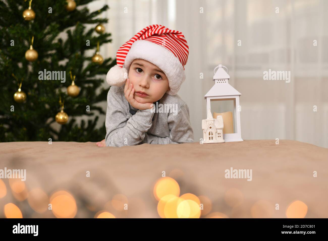 A boy with sad eyes, sitting with his hand holding his hand cheek. Dressed in cap. I leaned near the Christmas tree. Stock Photo