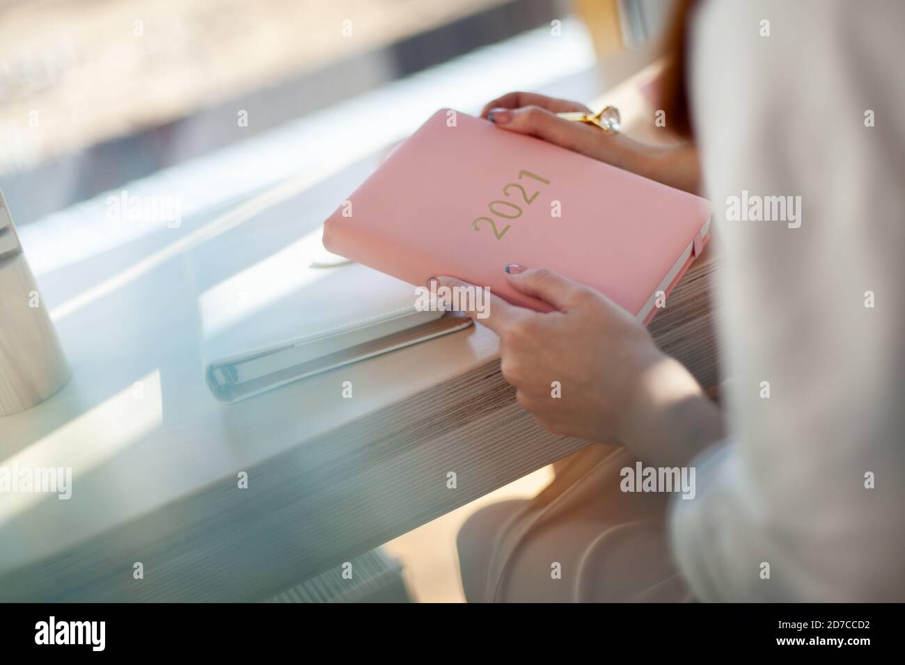 Female hands holding pink coral coloured leather diary 2021 while sitting near a window at cafe Stock Photo