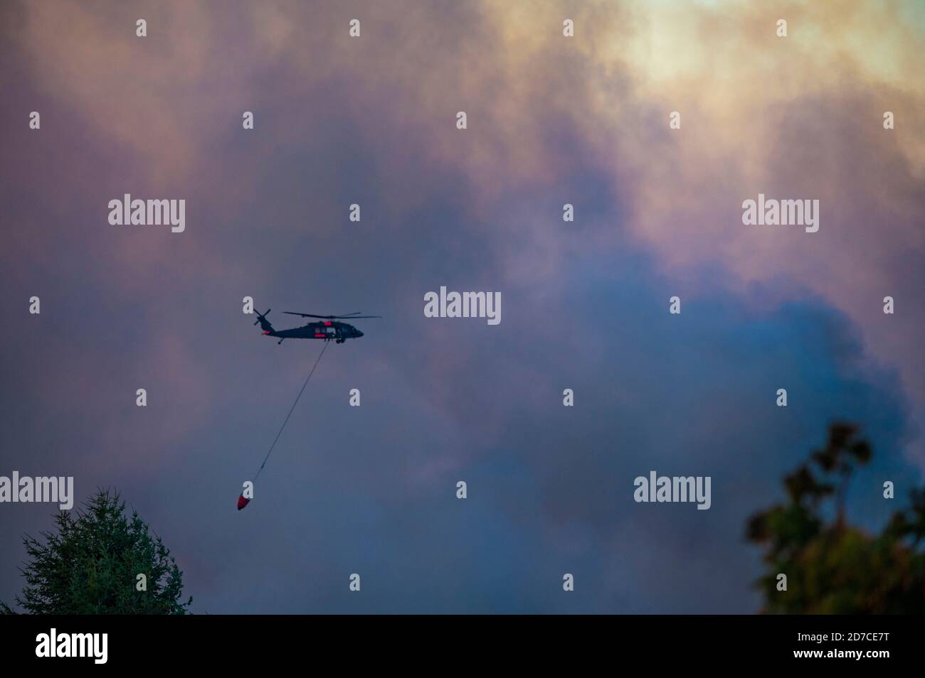 Aerial firefighting helicopter with fire bucket flying into smoke to drop water on wildfire Stock Photo