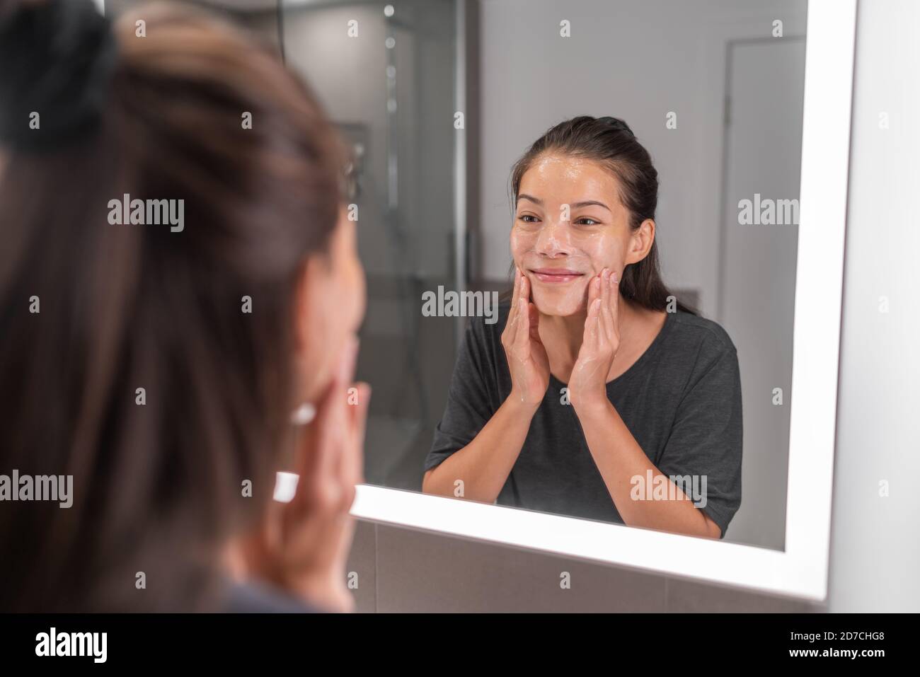 Woman washing face morning beauty skincare routing happy using soap foam in LED lighted mirror. Asian young girl massaging facewash exfoliate scrub Stock Photo