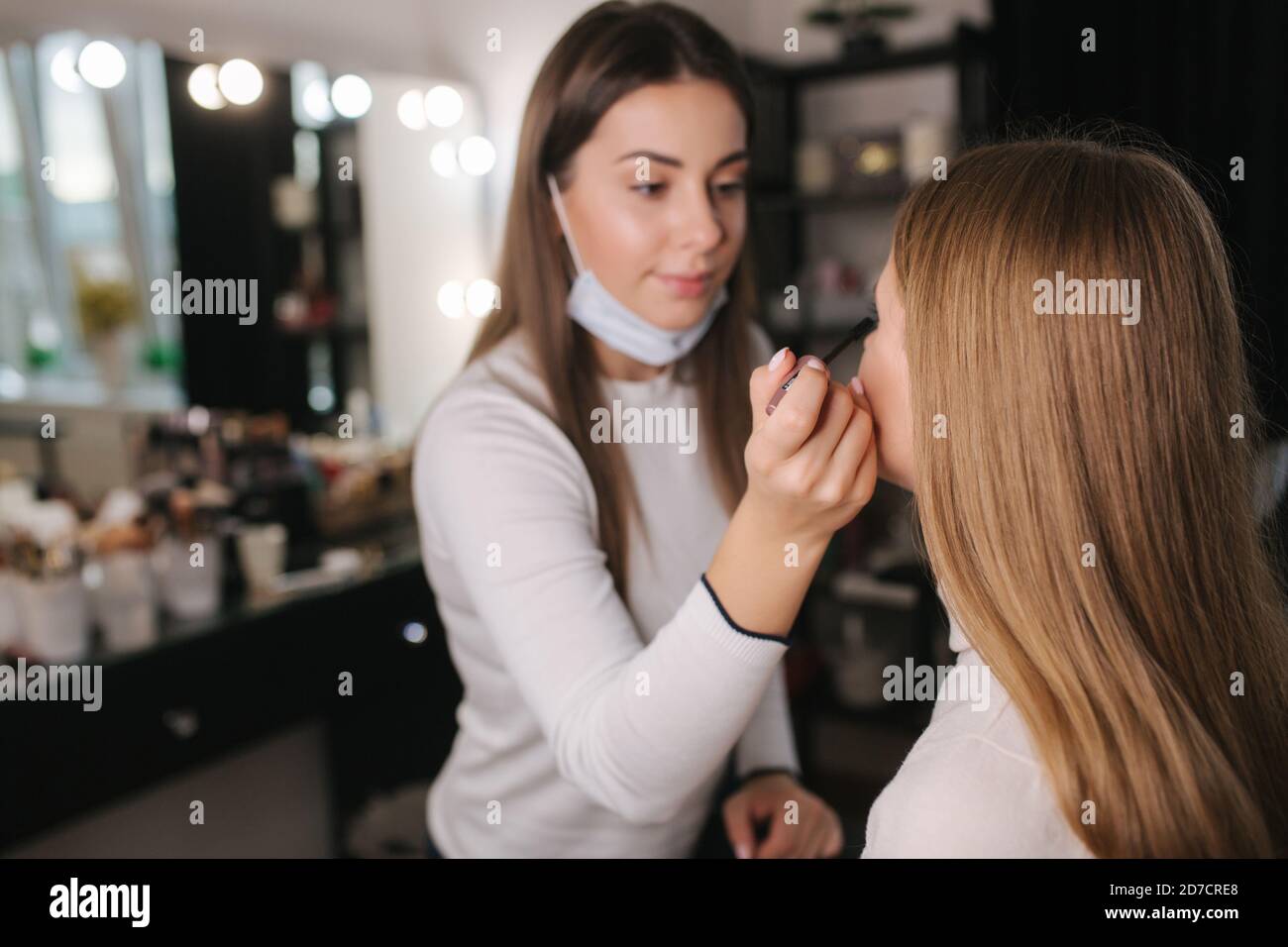 Female makeup artist working in beauty salon during quarantine. Make up artist in medical mask on chin. Beautiful blond hair model Stock Photo