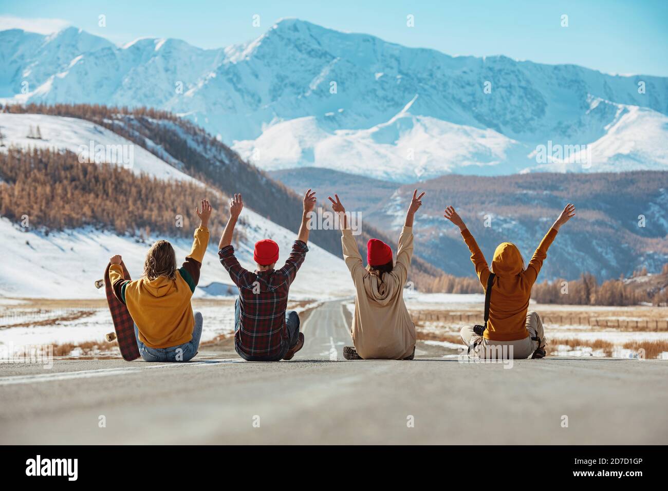 Four happy friends sits with raised arms on straight mountains road and having fun. Travel concept Stock Photo