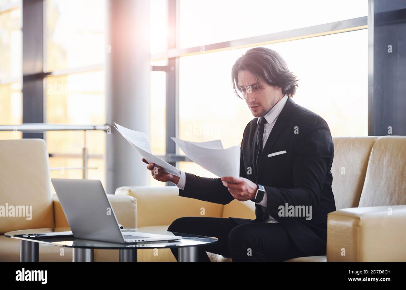 Sits on the sofa and reads documents. Portrait of handsome young businessman in black suit and tie Stock Photo