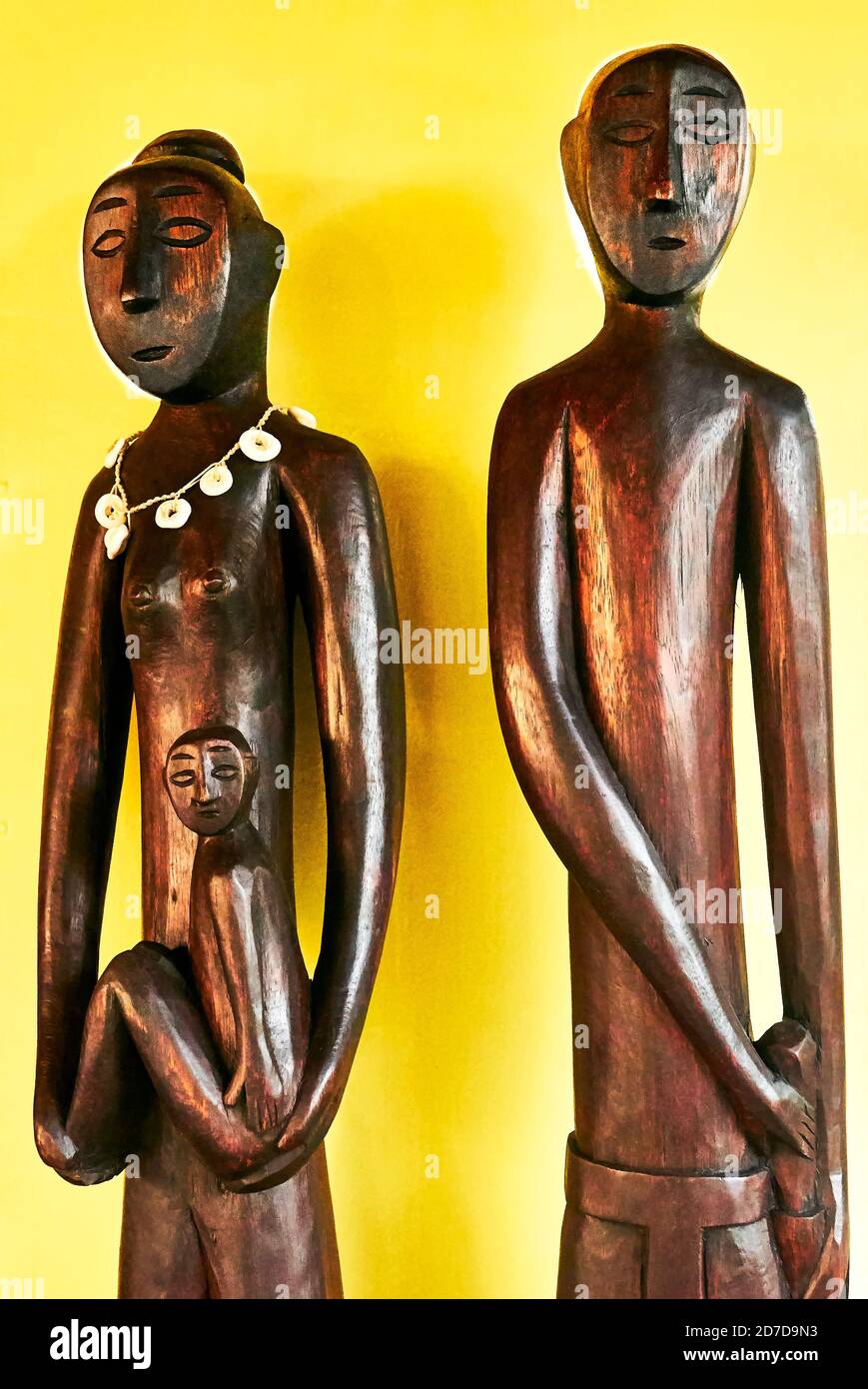 A mother holding her child next to the father as wooden carved statues standing in front of a yellow background, Puerto Princesa, Palawan, Philippines Stock Photo