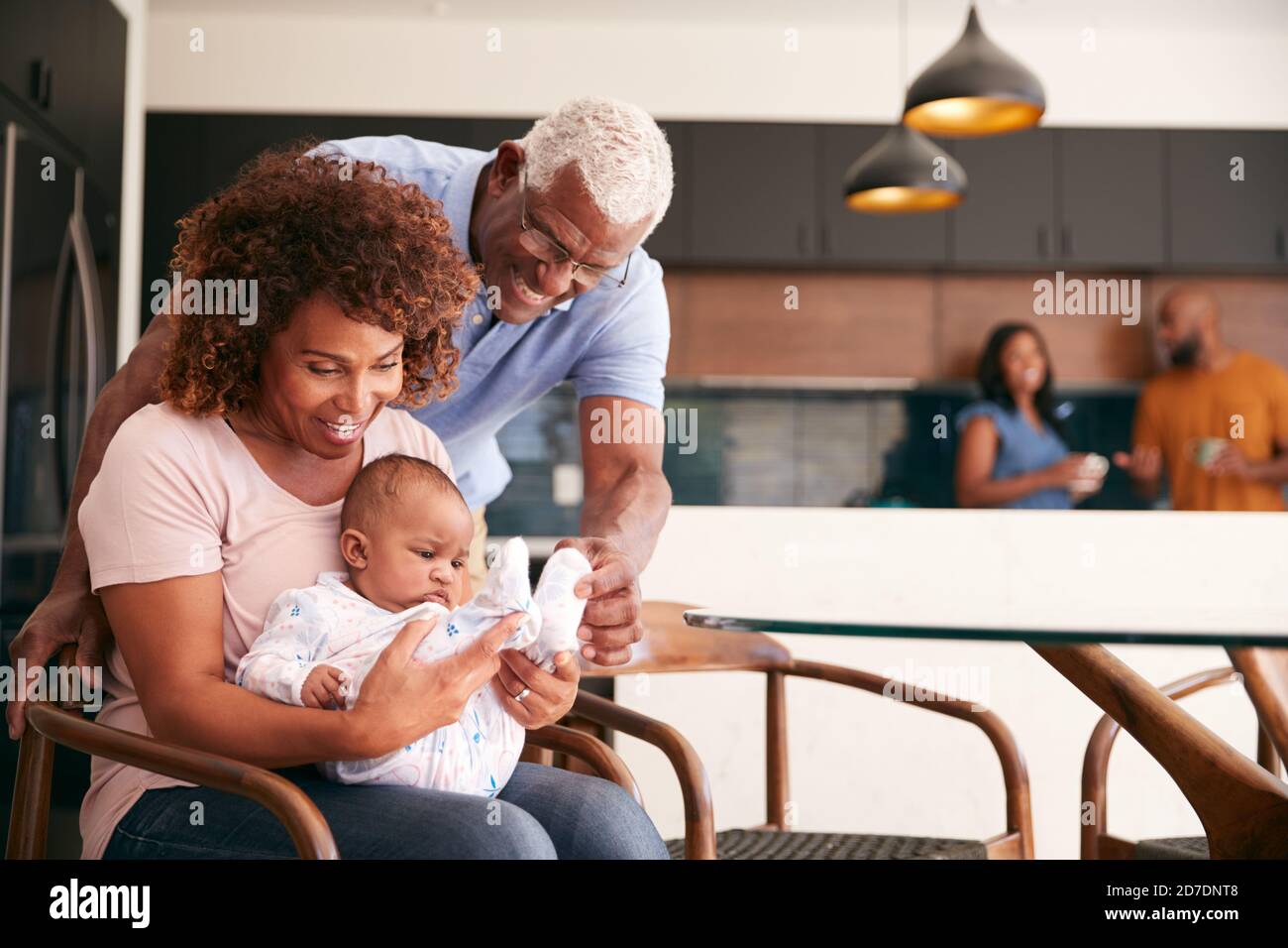 Grandparents Cuddling Baby Granddaughter At Home With Parents In Background Stock Photo
