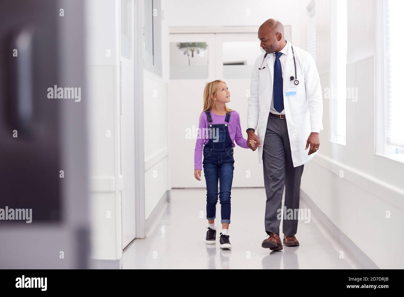 Male Paediatric Doctor Walking Along Hospital Corridor Holding Hands With Young Girl Patient Stock Photo