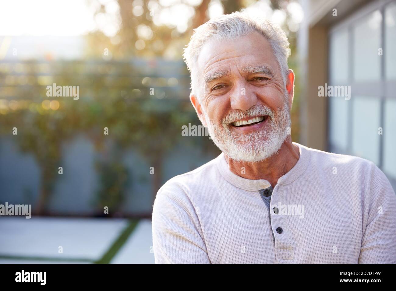 Portrait Of Smiling Retired Senior Hispanic Man In Garden At Home Against Flaring Sun Stock Photo