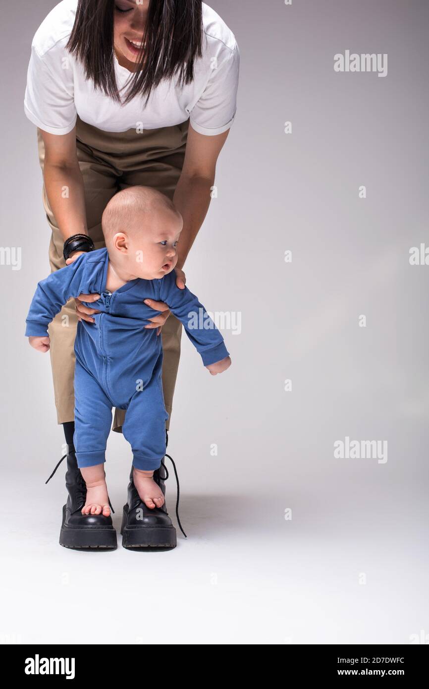 Mother encouraging baby kid To Take First Steps. A barefoot baby in a blue suit stands on mather's boots. Mom keeps safe  baby with his hands. Stock Photo