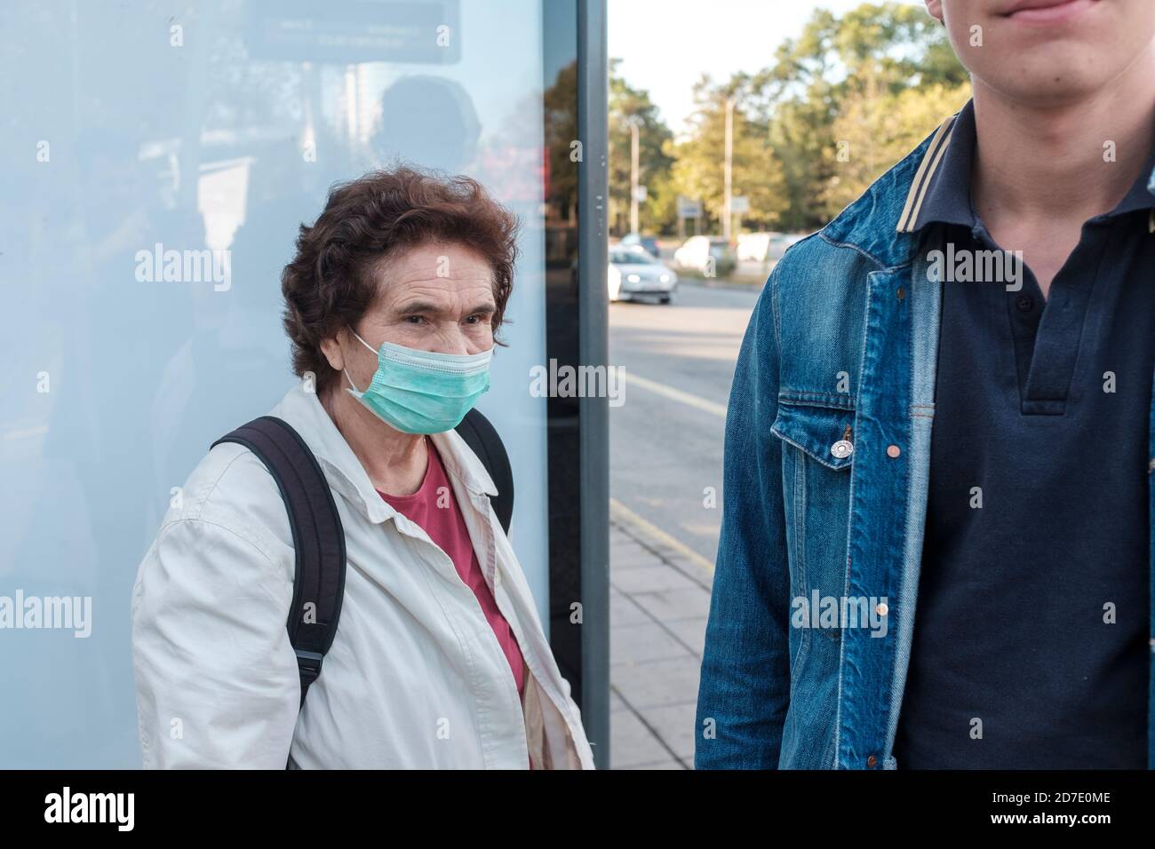 Covid 19-older woman wearing protective face mask next to young man without a mask at bus stop Stock Photo
