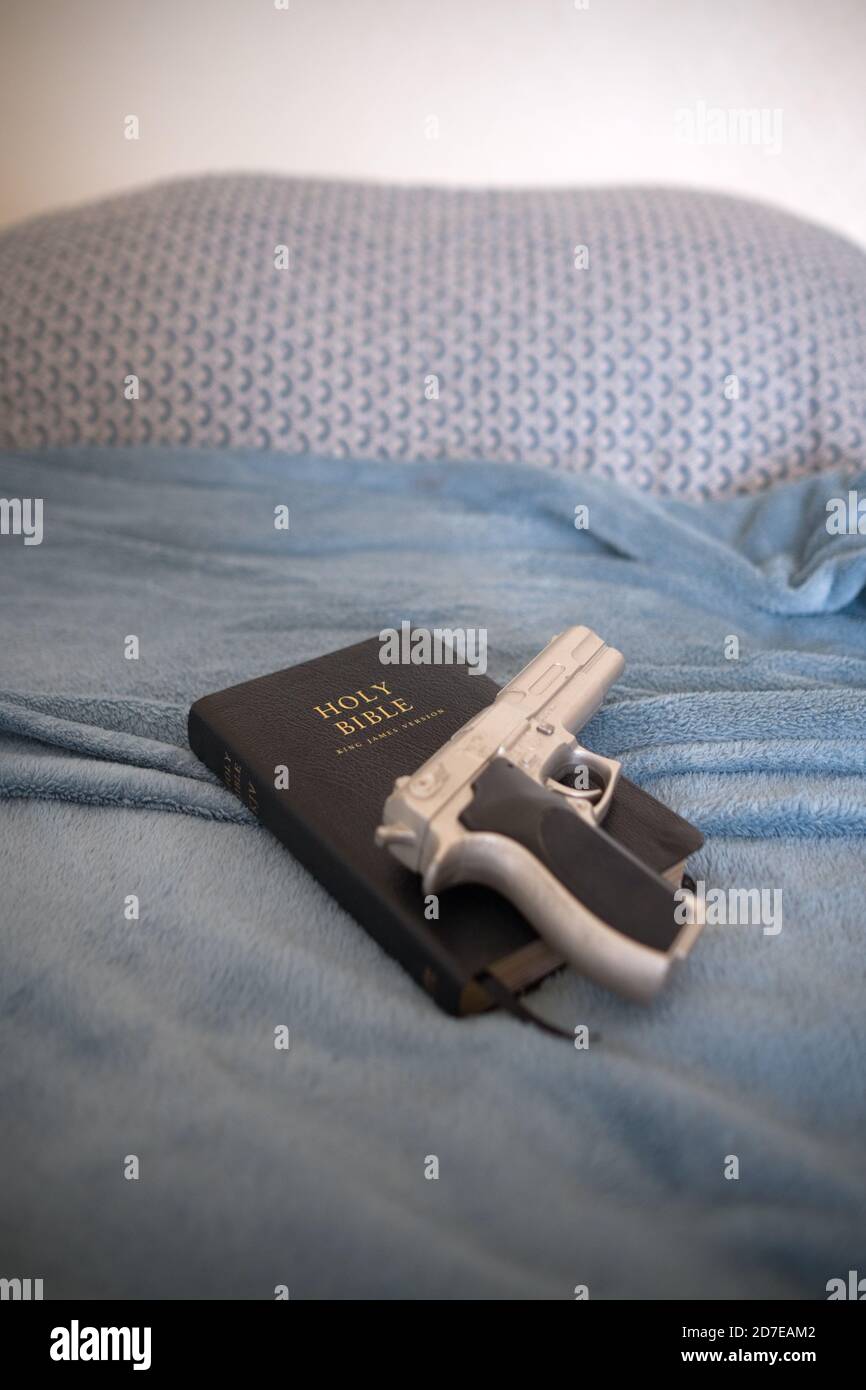 a silver replica berretta 9mm pistol on a black leather Holy Bible rests on a made bed in a bedroom in someones home Stock Photo